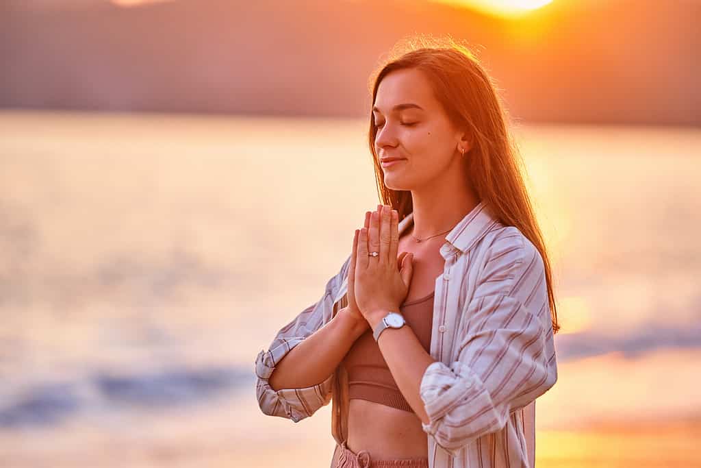 Portrait of calm serene satisfied woman with closed eyes and praying hands enjoys beautiful moment life on the seashore at sunset time